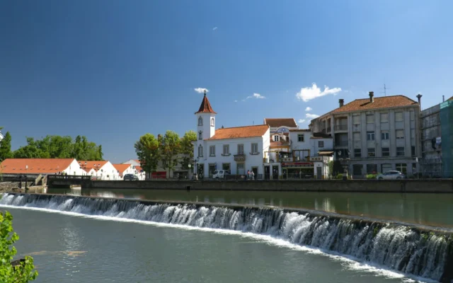 The Nabão river weir in Tomar, central Portugal, with white houses and a small bell tower on the far bank under a blue sky