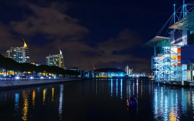Parque das Nações waterfront in Lisbon at night, with lit towers and buildings reflected in the water near the venue